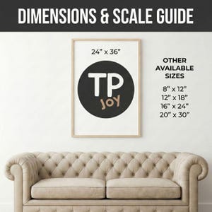 a beige tufted sofa against a white wall, with a framed poster or artwork hanging above it. The poster has a black and white design with the text "TPJOY" and dimensions provided.