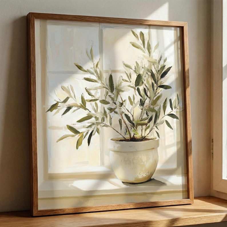 Framed olive branch wall art resting on a windowsill, softly lit by natural sunlight in a calm, neutral interior.