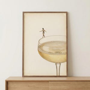 May include: A framed art print depicts a person balancing on the rim of a champagne glass filled with bubbly liquid. The artwork has a vintage aesthetic, with a warm, neutral background and a wooden frame. The image evokes a sense of surrealism and whimsy.
