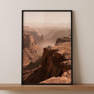 May include: Framed print of a vast canyon landscape in shades of brown and tan. The image captures a winding road and a group of people on a cliff edge. The sky is hazy, and the overall scene evokes a sense of depth and scale.