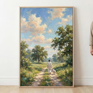 Op de afbeelding: Een ingelijst landschapsschilderij toont een figuur in witte gewaden die met een lam over een zandpad loopt. De scène speelt zich af in een veld met bomen onder een blauwe lucht vol wolken. Het kunstwerk zit in een houten lijst.