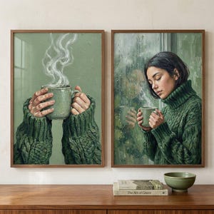 May include: Two framed art prints depict a cozy scene. One shows hands holding a green mug with steam rising, the other a person holding a mug by a window. Both wear green sweaters. The art is set on a wooden surface.