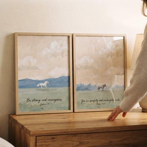 May include: Two framed wall art prints featuring watercolour landscapes with horses. The left print shows a white horse with the text Be strong and courageous. Joshua 1:9. The right print shows a brown horse with the text You are fearfully and wonderfully made. Psalm 139:14.