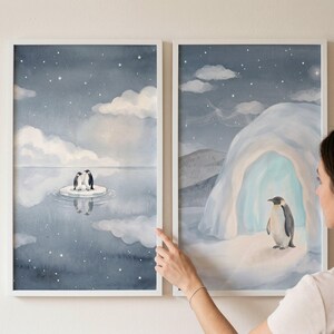 May include: Two framed watercolor prints featuring penguins in a snowy landscape. One print shows two penguins on an ice floe under a starry sky. The other print depicts a penguin near an ice cave. The prints have white frames.
