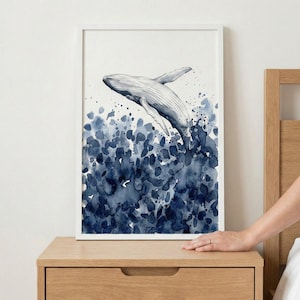 May include: Watercolor painting of a whale breaching the water. The artwork features a white whale against a backdrop of dark blue watercolor splashes. The painting is framed in a white rectangular frame.