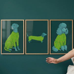 May include: Three framed art prints featuring stylized dog illustrations. Each dog is depicted in a different pose and wearing a green onesie with a rainbow pattern. The dogs are set against a dark teal background.