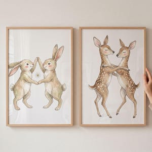 May include: Two framed watercolor prints. One features two rabbits holding hands with a daisy between them. The other print shows two deer embracing. Both prints have light brown wooden frames and are on a white background.