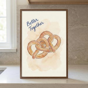 May include: A framed art print featuring two pretzels with sesame seeds, set against a watercolor wash. The words "Better Together" are written in navy blue script above the pretzels. The frame is a dark brown color.