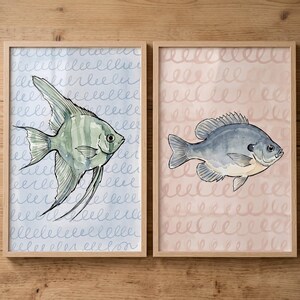 May include: Two framed wall art prints featuring watercolor fish illustrations. The left print shows a green angelfish on a light blue background with a repeating swirl pattern. The right print displays a blue fish on a soft pink background.