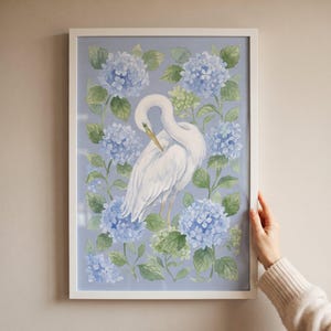 May include: A framed vertical wall art print featuring a white egret bird with a yellow beak standing amidst clusters of light blue hydrangea flowers and green leaves against a soft periwinkle blue background, enclosed in a white frame.