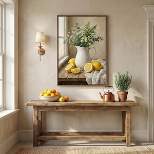 An interior scene featuring a rustic wooden console table against a neutral-toned wall. Centered above the table is a large framed vertical artwork of a still life with a white pitcher of daisies and greenery, surrounded by lemons. On the table, a bo
