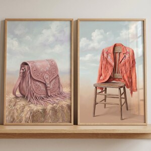 May include: Two framed wall art prints featuring a dusty pink leather saddle bag with fringe on a hay bale and an orange suede jacket with fringe draped over a wooden chair, both set against a soft blue sky with white clouds background.