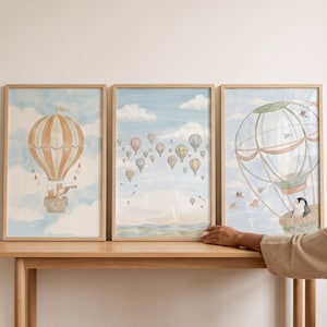 May include: Three framed art prints featuring hot air balloons in a whimsical style. The prints showcase a fox in a balloon, a penguin in a balloon, and a sky filled with balloons. The color palette includes soft blues, oranges, and browns.