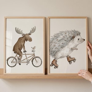 May include: Two framed watercolor prints. One features a moose riding a bicycle with a bird perched on the handlebars. The other print shows a hedgehog wearing roller skates. Both prints have a light beige background and are in wooden frames.