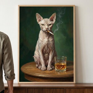 A framed artwork depicting a hairless Sphynx cat with a stern expression sitting on a round wooden table. The cat holds a lit cigarette in its mouth, with wisps of smoke curling upwards. To its side sits a crystal glass filled with an amber-colored l