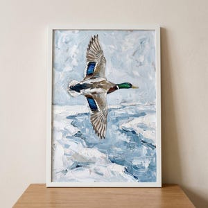 Textured winter mallard duck wall art depicts a bird in flight over icy blue water, displayed in a white frame.