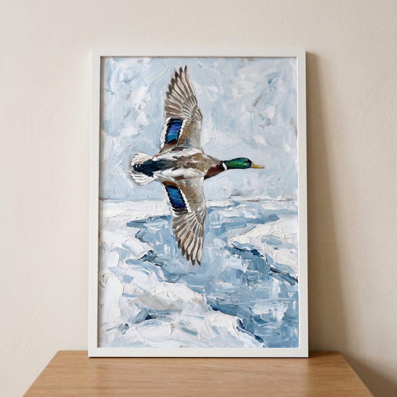 Textured winter mallard duck wall art depicts a bird in flight over icy blue water, displayed in a white frame.