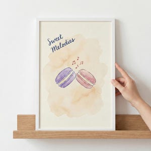 Könnte beinhalten: Gerahmter Aquarelldruck mit zwei Makronen, einer lila und einer rosa, mit Noten und dem Text "Sweet Melodies". Die Makronen sind vor einem pfirsichfarbenen Aquarellhintergrund in einem weißen Rahmen.