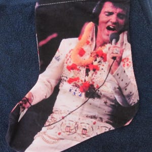 May include: A Christmas stocking featuring a printed image of Elvis Presley performing in a white jumpsuit with a microphone in hand, adorned with a yellow and red lei around the neck, and a small red hanging loop at the top corner.