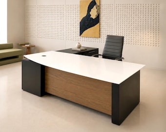 Puede incluir: Un escritorio ejecutivo grande con una superficie rectangular blanca, un panel central con veta de madera y soportes laterales negros. Una silla de oficina negra con costuras horizontales se sitúa detrás del escritorio, que incluye un ala y un cajón.