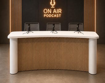 Puede incluir: Un escritorio de podcast curvo con superficie blanca y frente de listones de madera clara, flanqueado por dos pilares cilíndricos blancos. Tres micrófonos negros sobre soportes descansan en el escritorio. Un letrero de neón dice ON AIR PODCAST.