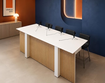 Puede incluir: Una mesa rectangular blanca larga con una base de listones de madera clara y soportes verticales blancos. Cuatro taburetes negros se sitúan a lo largo de la mesa, que tiene cuatro micrófonos negros en trípodes conectados por cables.