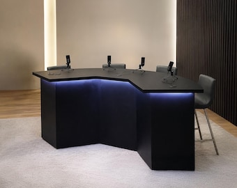 Puede incluir: Un escritorio de podcast negro en forma de V con cuatro micrófonos montados en trípodes pequeños, iluminación LED azul integrada a lo largo del borde frontal y cuatro taburetes de bar grises tapizados con patas de metal alrededor.