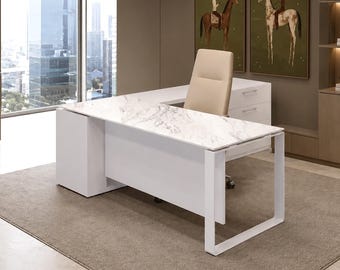 Puede incluir: Un escritorio en forma de L con una superficie de patrón de mármol blanco, una estructura blanca sólida y una unidad de almacenamiento lateral con cajones. Una silla de oficina beige de respaldo alto está frente al escritorio sobre una alfombra gris.