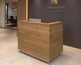 Puede incluir: Un mostrador de recepción de madera marrón claro con base plateada se sitúa frente a una pared gris oscuro con listones verticales y el logotipo y texto metálicos Northridge Solutions. Un monitor plateado es visible sobre el mostrador.