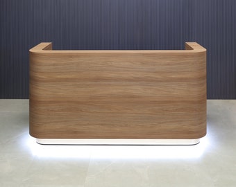 Puede incluir: Un mostrador de recepción rectangular con un acabado de veta de madera marrón claro y bordes laterales redondeados. La base del mostrador está elevada por una tira de luz blanca que ilumina el suelo directamente bajo la estructura.