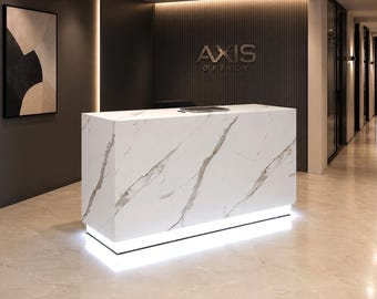 Puede incluir: Un mostrador de recepción rectangular con superficie de mármol blanco y vetas grises, con una base iluminada por luces LED integradas. El mostrador se sitúa frente a una pared gris oscuro con el texto metálico AXIS OFFICE montado.