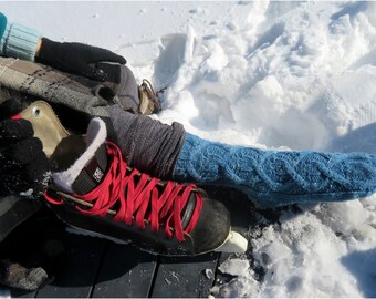 May include: A pair of black ice skates with red laces. The skates are on a wooden surface with snow in the background. One skate is being put on by a person wearing blue knitted socks.