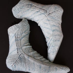 May include: A pair of light blue knitted socks with a lacy pattern. The socks are made with a yarn that has a subtle marled effect.