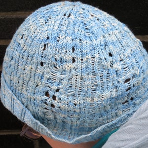 A woman wearing a lace-and cable beanie hat with an intricate cable stitch. The hat is knit in a pale-blue fingering-weight yarn.
