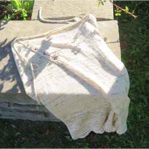 May include: Cream-colored knitted shawl with a scalloped edge and small red accents. The shawl is made of a soft, textured yarn and has a drawstring closure. The shawl is draped over a concrete surface in natural sunlight.