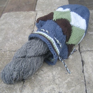 This shows a drawstring bag made primarily of joined mitered squares. The top has eyelets with a twisted cord threaded through them, and a skein of yarn is partially out of the open bag.