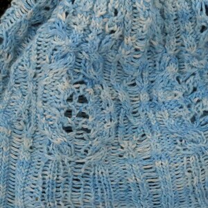 A lace hat laying flat. The lace and cable pattern (eyelets, loop stitches and two-stitch cables) runs vertically along the hat and meets at the crown of the hat. Hat is knit in pale blue fingering-weight yarn.