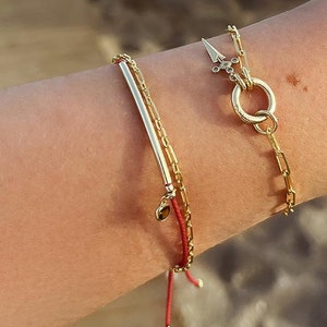 May include: A close-up of a person's wrist wearing three gold bracelets. The bracelets are layered on top of each other. The top bracelet is a chain with a large, round charm. The middle bracelet is a thin, gold bar. The bottom bracelet is a red string with a small gold charm.