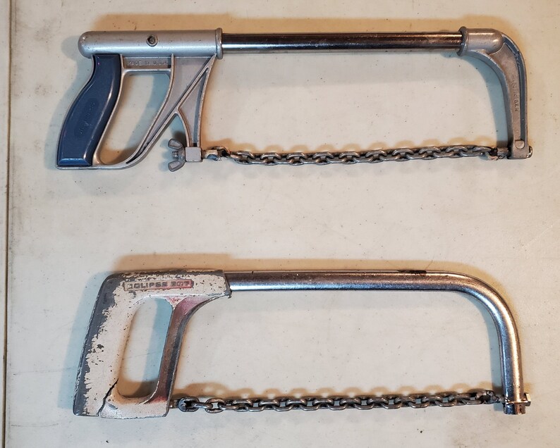 May include: Two vintage hand saws with chains. The top saw has a blue handle and the bottom saw has a white handle with the text "Eclipse 307" printed on it.