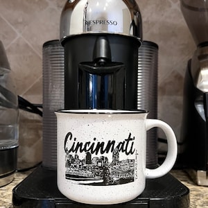 May include: White ceramic mug with a black rim and black speckles. The mug has a black and white illustration of the Cincinnati skyline with the word "Cincinnati" in a black cursive font. The mug is sitting on a black Breville coffee machine.