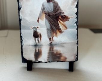 May include: A rectangular slate plaque featuring a religious image of a figure in white robes walking with a lamb in shallow water. The image has a serene, soft color palette with a black stand.