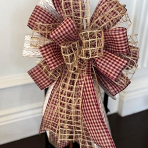 Puede incluir: Un lazo decorativo con capas de cinta de cuadros burdeos y blancos, acentuado con una red de tono natural y una cinta estampada color crema. Diseñado para la decoración de temporada o festiva.