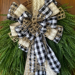 May include: A decorative wreath featuring a large bow with black and white checkered ribbon, tan burlap ribbon, and green foliage. The bow is the focal point, with multiple layers and textures. The wreath is suitable for seasonal home decor.