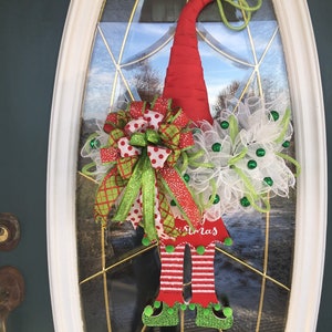 May include: A Christmas gnome wreath with a red hat, green and white bow, and green and white mesh. The gnome's legs are made of wood and have the word "Christmas" written on them.