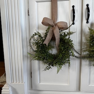 May include: A green fern wreath with a brown burlap bow hangs on a white cabinet door. The wreath is round and full, with small green leaves. The bow is large and decorative, adding a rustic touch to the decor.