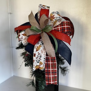 May include: A decorative fall-themed bow arrangement featuring a variety of ribbons in red, navy, brown, and white with leaf and plaid patterns. The bow is atop a black rectangular base with faux greenery.