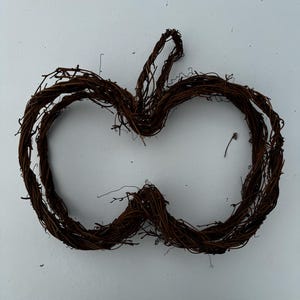 May include: A rustic, brown grapevine wreath in the shape of an apple. The wreath is made of intertwined twigs and branches, creating a natural, textured appearance. The apple shape is complete with a stem at the top, perfect for fall decor.