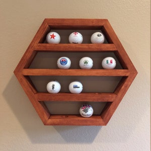 May include: A hexagonal wooden shelf with six shelves, each holding a golf ball with a different design. The shelf is stained a dark reddish-brown color and is mounted on a wall.