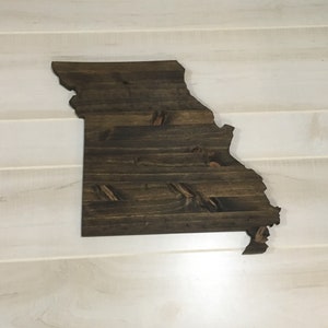 May include: A wooden wall hanging in the shape of the state of Missouri. The wood has a dark brown stain, with visible wood grain and knots. The piece is mounted on a white, horizontal-slatted wall.