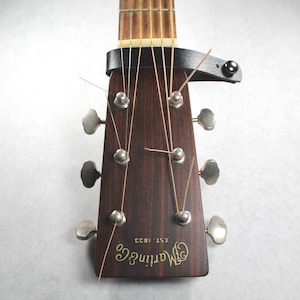May include: Close-up of a guitar headstock with a black capo. The headstock is dark brown with silver tuning pegs and strings. The brand name "Martin & Co. EST. 1833" is visible.